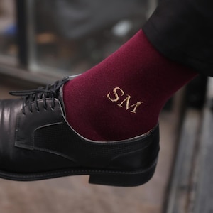 Può includere: Calzini bordeaux con iniziali "SM" ricamate in oro, visibili sopra una scarpa in pelle nera. I calzini sono tirati su, mostrando le iniziali. La scarpa è di stile classico.