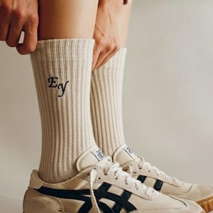 May include: Cream-colored athletic socks with the embroidered letters "Ey" in navy blue are pulled up over the ankles. The socks are worn with off-white sneakers with navy blue stripes and a brown sole.