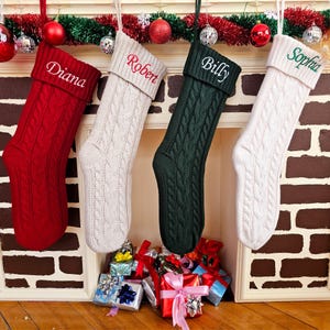May include: Four cable-knit Christmas stockings in red, cream, dark green, and white hang from a mantel. Each stocking is personalized with a name embroidered in a contrasting color: "Diana," "Robert," "Billy," and "Sophia." Presents are piled below.