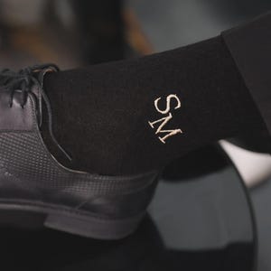 Puede incluir: Calcetines negros con las iniciales "SM" bordadas en dorado claro. Los calcetines se llevan con un zapato de cuero negro. El zapato tiene un patrón texturizado en la parte superior.