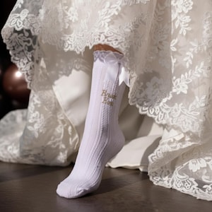 May include: White ribbed knit sock with the words "Bride Tribe" embroidered in gold. The sock has a white bow at the top and is partially visible under a white lace wedding dress.