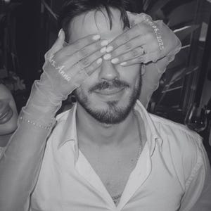 May include: A monochrome image of a man in a white shirt, with a beard. His eyes are covered by sheer gloves embroidered with "Mrs. Anna" and "01.06.2025". The gloves are held by a person.