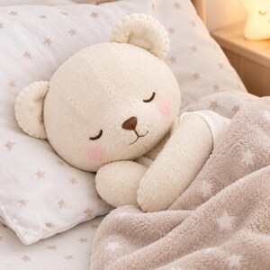May include: A plush cream-colored teddy bear with closed eyes and pink cheeks, resting its head on a white pillow patterned with small beige stars. The bear is partially covered by a soft, fuzzy beige blanket featuring a white star pattern.