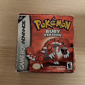 May include: A Game Boy Advance video game box for Pokémon Ruby Version. The box is red with the Pokémon logo and a graphic of a red and white creature. The box also has the Game Boy Advance logo and the Nintendo logo.