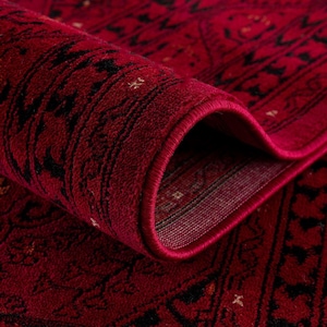 Op de afbeelding: Close-up van een opgerolde, rijk gestructureerde rode vloerkleed met ingewikkelde zwarte patronen. Het ontwerp van het vloerkleed heeft een gedetailleerde rand en herhalende motieven, die een traditionele esthetiek laten zien. De opgerolde rand onthult de dikte en constructie van het vloerkleed.