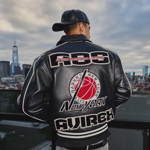 Puede incluir: Una chaqueta de cuero negra con texto y gr&aacute;ficos bordados en blanco y rojo en la espalda. El dise&ntilde;o incluye el texto "ABS", "AVIREX BASKETBALL SPORTS", "NEW YORK" y "AVIREX" con un gr&aacute;fico de baloncesto rojo central. Se lleva una gorra negra.