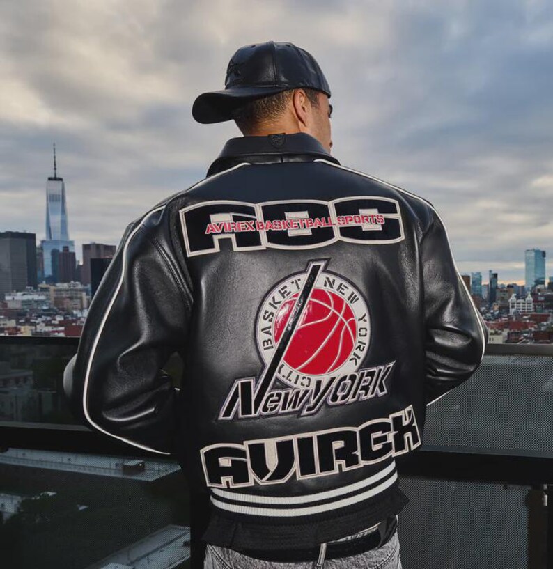 Puede incluir: Una chaqueta de cuero negra con texto y gr&aacute;ficos bordados en blanco y rojo en la espalda. El dise&ntilde;o incluye el texto "ABS", "AVIREX BASKETBALL SPORTS", "NEW YORK" y "AVIREX" con un gr&aacute;fico de baloncesto rojo central. Se lleva una gorra negra.