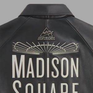 May include: Black leather jacket with white embroidered text that reads "MADISON SQUARE" and the "AVIREX" logo. The jacket has a classic collar and a stitched design on the back.