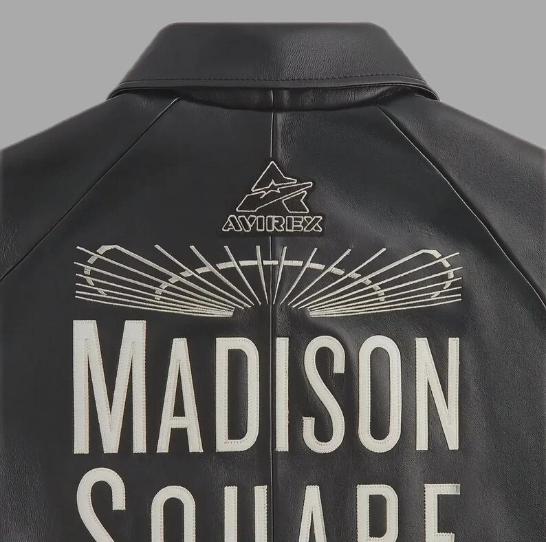 May include: Black leather jacket with white embroidered text that reads "MADISON SQUARE" and the "AVIREX" logo. The jacket has a classic collar and a stitched design on the back.