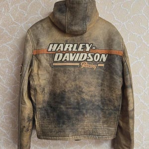 Op de afbeelding: Een bruine leren jas met een distressed look en een capuchon. De achterkant heeft het Harley-Davidson Racing logo in wit en oranje. De jas heeft een verweerde uitstraling, wat wijst op een vintage stijl.