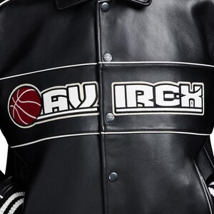 Puede incluir: Una chaqueta universitaria de cuero negro con una franja horizontal blanca en el pecho con el texto MAV IRCH en letras may&uacute;sculas blancas. Un gr&aacute;fico circular de un bal&oacute;n de baloncesto rojo se sit&uacute;a a la izquierda del texto. Botones de presi&oacute;n plateados.