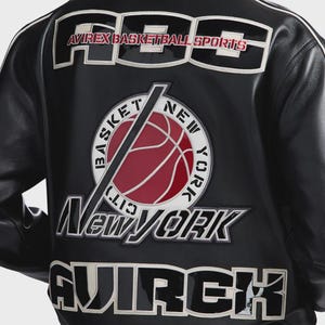 Puede incluir: Una chaqueta de cuero negra con grandes letras bordadas en blanco y negro que dicen ABS y AVIREX. Un parche circular en el centro muestra un bal&oacute;n de baloncesto rojo con el texto BASKET NEW YORK CITY en blanco alrededor.