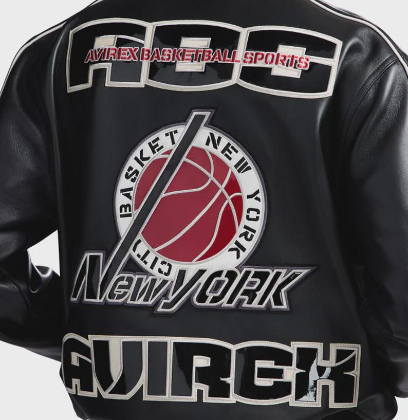 Puede incluir: Una chaqueta de cuero negra con grandes letras bordadas en blanco y negro que dicen ABS y AVIREX. Un parche circular en el centro muestra un bal&oacute;n de baloncesto rojo con el texto BASKET NEW YORK CITY en blanco alrededor.