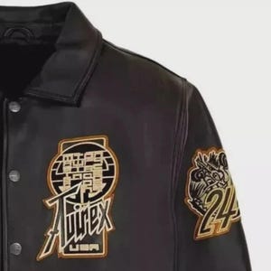 以下が含まれることがあります： 黒い革のジャケットで、ボタン留めと襟が付いています。 ジャケットには、「Aviirex USA」の文字と数字の「24」が入ったドラゴンなど、金と白のアクセントが付いた刺繍パッチが付いています。