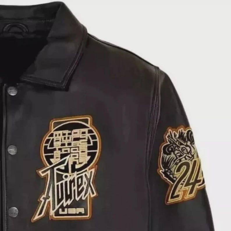 以下が含まれることがあります： 黒い革のジャケットで、ボタン留めと襟が付いています。 ジャケットには、「Aviirex USA」の文字と数字の「24」が入ったドラゴンなど、金と白のアクセントが付いた刺繍パッチが付いています。