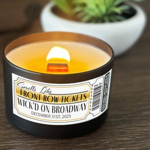 May include: A black candle with a wooden wick and a label that reads "Smells Like Front Row Tickets Wick'd on Broadway December 31st, 2025." The candle has a golden wax and is in a black container.
