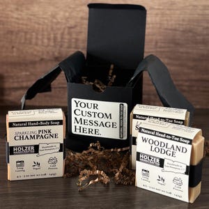 May include: Two rectangular bars of Holzer Handcrafted soap in Sparkling Pink Champagne and Woodland Lodge scents, each weighing 4.5-5 oz, displayed with a black gift box featuring a white label that reads Your Custom Message Here.