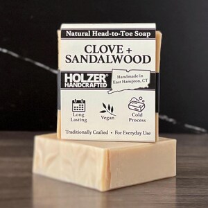 May include: A rectangular bar of light beige soap wrapped in a white paper label. The label reads Natural Head-to-Toe Soap, CLOVE + SANDALWOOD, HOLZER HANDCRAFTED, Handmade in East Hampton, CT, Long Lasting, Vegan, Cold Process, Traditionally Crafted.