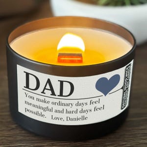 May include: A lit black candle with a wooden wick, labeled "DAD" with a blue heart. The label includes the text: "You make ordinary days feel meaningful and hard days feel possible. Love, Danielle."