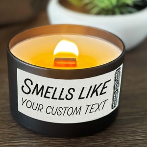 May include: A lit black candle with a wooden wick, featuring the text "Smells Like Your Custom Text" on a white label. The candle has a warm, orange glow and is set in a dark container. A small label on the side reads "Natural Wood Wick Coconut Soy Candle."