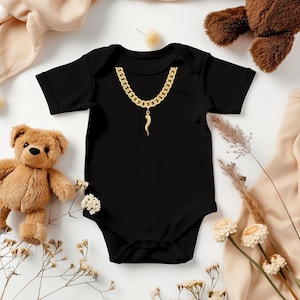 May include: Black baby bodysuit with a gold chain and pendant design. The short-sleeved onesie features a gold chain necklace graphic with a pendant. The baby garment is laid flat on a white surface with teddy bears and floral accents.