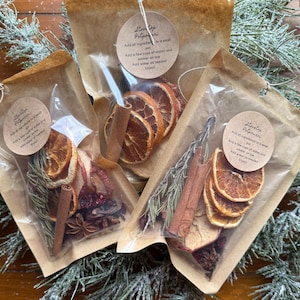 Könnte beinhalten: Drei Beutel mit Stovetop-Potpourri, die jeweils getrocknete Orangenscheiben, Zimtstangen und andere aromatische Zutaten enthalten. Die Beutel sind in klarem Kunststoff versiegelt und mit Bindfaden gebunden, mit einem runden Anhänger.