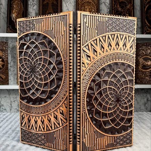 Könnte beinhalten: Ein zweiteiliger Holzschirm mit aufwendigen geometrischen Mustern. Der Schirm ist hauptsächlich braun mit goldenen Akzenten und weist ein zentrales kreisförmiges Design und lineare Elemente auf. Die Paneele sind durch Scharniere verbunden.