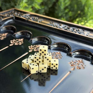 Könnte beinhalten: Ein Stapel aus acht cremefarbenen sechsseitigen Würfeln mit schwarzen Augen, die auf einem glänzend schwarzen Backgammon-Spielbrett mit goldenen dekorativen Ornamenten und bogenförmigen Vertiefungen liegen.
