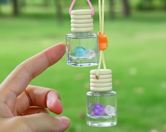 May include: Two small, clear glass bottles with wooden tops and colorful beads inside are suspended by strings. One bottle has a blue bead, the other a purple bead. An orange clasp connects the strings. The background is a blurred green.