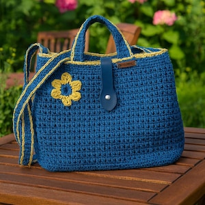 Può includere: Borsa a mano blu all'uncinetto fatta a mano con un'applicazione floreale gialla. La borsa presenta finiture e cinturino gialli abbinati e una chiusura in pelle blu. La borsa è appoggiata su un tavolo di legno.