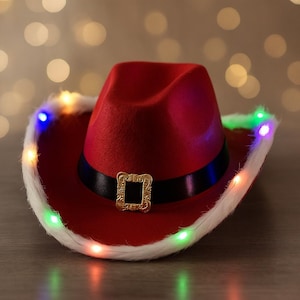 Puede incluir: Un sombrero de vaquero rojo con una banda negra y hebilla dorada. El borde del sombrero está adornado con piel sintética blanca y luces LED de colores. Un accesorio festivo.
