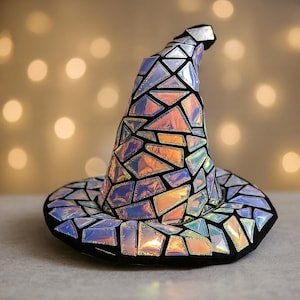 Puede incluir: Un sombrero de bruja caprichoso con un diseño de mosaico. El sombrero presenta formas geométricas iridiscentes en tonos naranja, azul y plata, delineadas en negro. El ala y la punta del sombrero también son negras.