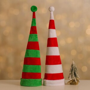 May include: Two plush Christmas elf hats. One is green and red striped with a green ball on top. The other is red and white striped with a white ball on top. A small decorative Christmas tree is also present.