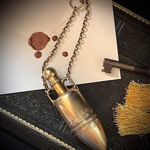 May include: A gold-toned metal bottle with an ornate design and a chain attached to a ring. The bottle is sitting on a dark, textured surface.