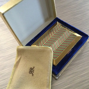 May include: A gold and blue compact with a geometric pattern on the lid. The compact is open and has a beige velvet lining with the initials "RR" embroidered in the center.