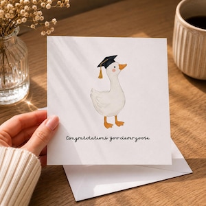 Può includere: Un biglietto d'auguri quadrato bianco con un'illustrazione ad acquerello di un'oca bianca che indossa un tocco di laurea nero con una nappa dorata. Sotto l'oca, il testo recita "Congratulations you clever goose" in un semplice carattere nero.