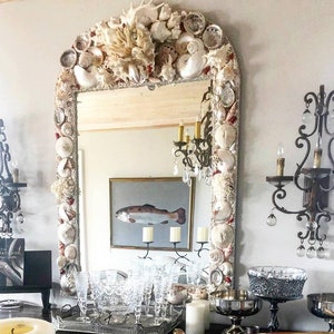 May include: An arched mirror framed with various seashells and coral. The mirror reflects a painting of a fish and a chandelier. Decorative sconces flank the mirror, and a table displays glassware and candles.