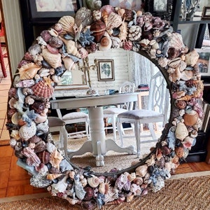 May include: A circular mirror with a dark frame, adorned with an array of seashells in white, beige, brown, and blue hues. The mirror reflects a room with a table and chairs, evoking a coastal or nautical aesthetic.