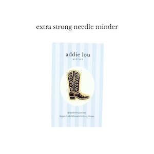 Könnte beinhalten: Ein Nadelhalter mit einem braunen und goldenen Cowboy-Stiefel-Design. Der Stiefel befindet sich vor einem hellblau gestreiften Hintergrund mit dem Text "addielou atelier". Der Text "extra strong needle minder" ist ebenfalls vorhanden.