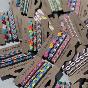 Puede incluir: Una colección de pulseras de la amistad coloridas. Las pulseras presentan varios patrones tejidos y combinaciones de colores, incluyendo azul, rosa, amarillo y verde. Cada pulsera está unida a una tarjeta marrón con el texto "BENNY + LUKA" y "Handmade with love."