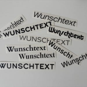 Könnte beinhalten: Eine Ansammlung weißer, rechteckiger Aufkleber mit dem schwarzen Aufdruck "Wunschtext". Die Aufkleber liegen auf einer weißen Oberfläche und sind unterschiedlich ausgerichtet.