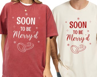 Op de afbeelding: Twee t-shirts, één rood en één crème, elk met de tekst "SOON TO BE Merry'd" in wit en rood, samen met een afbeelding van twee in elkaar grijpende ringen. De shirts zijn waarschijnlijk voor een stel.