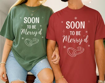Op de afbeelding: Twee shirts met korte mouwen, één groen en één rood, met de tekst "SOON TO BE Merry'd" in het wit. Het ontwerp bevat sneeuwvlokken en ringafbeeldingen. Het paar draagt ​​een blauwe jeans.