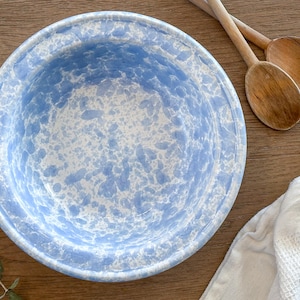 May include: A round, blue and white speckled enamelware bowl. Two wooden spoons and a white cloth are next to the bowl on a wooden surface. The bowl has a wide rim and a speckled pattern.