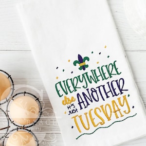 May include: White kitchen towel with embroidered text in green, purple, and gold. The text reads "EVERYWHERE else HAS JUST ANOTHER TUESDAY." A fleur-de-lis and small dots are also embroidered on the towel.