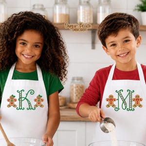 May include: Two white aprons featuring embroidered gingerbread men and large green letters K and M. The aprons are worn over a green shirt and a red long-sleeve shirt. The text Sugar Beach Embroidery is visible on a wooden shelf in the background.
