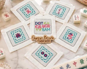 May include: A set of five square white fabric coasters featuring embroidered mahjong tile designs in teal and pink, with a central coaster displaying the text DOT CRACK BAM and a wooden Sugar Beach Embroidery logo sign.
