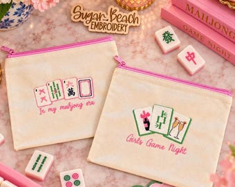 May include: Two off-white canvas zipper pouches with pink trim. The left pouch features embroidered mahjong tiles and the text In my mahjong era. The right pouch displays embroidered mahjong tiles with a bottle and glasses and the text Girls Game Night.