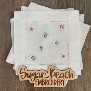 May include: A stack of white linen cocktail napkins featuring embroidered mahjong tile motifs in various colors. A wooden sign with the laser-cut text Sugar Beach EMBROIDERY is placed in front of the napkins on a wooden surface.