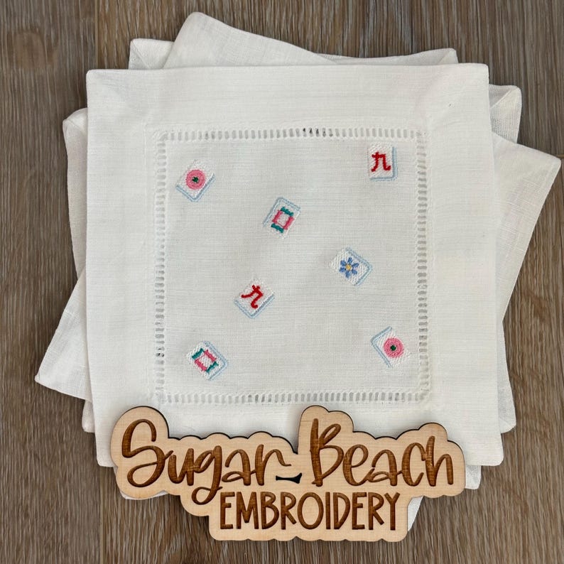 May include: A stack of white linen cocktail napkins featuring embroidered mahjong tile motifs in various colors. A wooden sign with the laser-cut text Sugar Beach EMBROIDERY is placed in front of the napkins on a wooden surface.